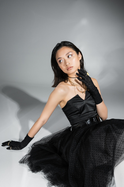 chic asian young woman with short hair sitting in black strapless dress with tulle skirt with belt and gloves while looking away and touching neck on grey background, wet hairstyle, golden necklaces  - Foto, Imagem