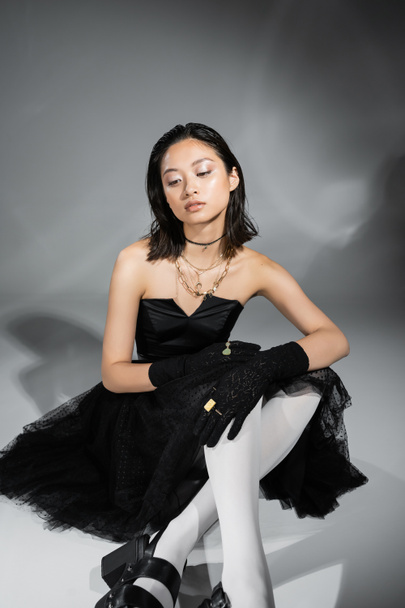 rêveuse asiatique jeune femme aux cheveux courts assis en robe bustier noire avec jupe en tulle collants blancs, chaussures et gants tout en regardant loin sur fond gris, coiffure humide, colliers dorés  - Photo, image