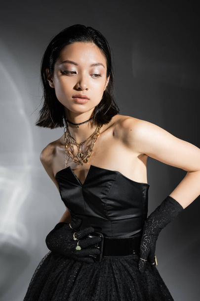 charming asian young woman with short hair posing in black strapless dress with tulle skirt with belt and gloves while looking away on grey background, wet hairstyle, golden jewelry  - Φωτογραφία, εικόνα
