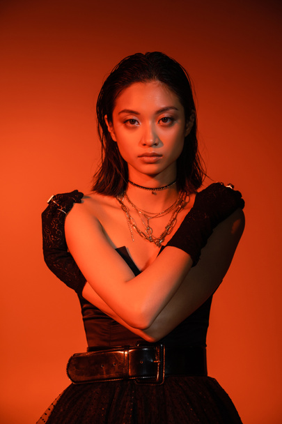 beautiful asian young woman with short hair and wet hairstyle posing with crossed arms in black strapless dress and gloves while standing on orange background with red lighting, golden jewelry  - Foto, afbeelding