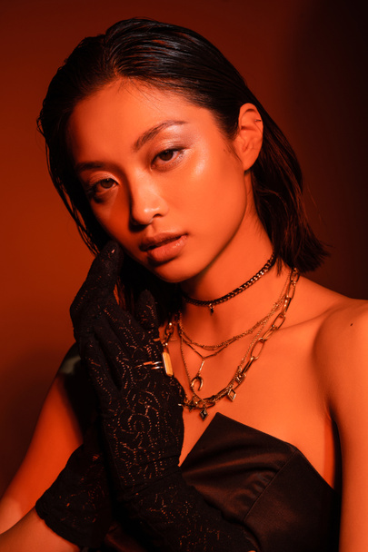 portrait of alluring asian young woman with short hair and wet hairstyle posing in black strapless dress and gloves while standing on orange background with red lighting, golden jewelry  - Foto, afbeelding