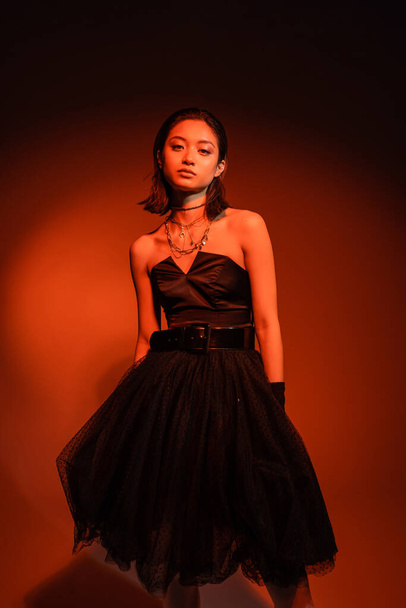 stylish asian woman with short hair and wet hairstyle posing in black strapless dress with tulle skirt and gloves while standing on orange background with red lighting, golden jewelry, young model - Photo, Image