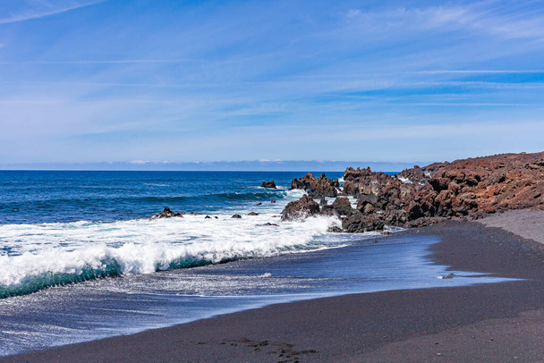 Bright contrast of white sea foam of the waves rolling onto shore with the sand and black solidified lava. - Photo, image