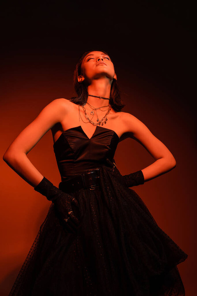 stylish asian woman with closed eyes and wet hairstyle posing with hands on hips in black strapless dress with tulle skirt and gloves while standing on orange background with red lighting, young model - Photo, Image