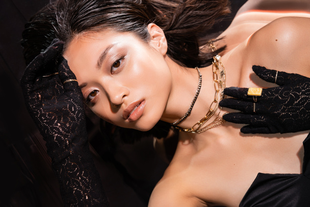 Draufsicht der schönen asiatischen Frau mit kurzen Haaren und nasser Frisur posiert in schwarzen Handschuhen mit goldenen Ringen, während sie auf dunklem Hintergrund in die Kamera schaut, junges Model  - Foto, Bild