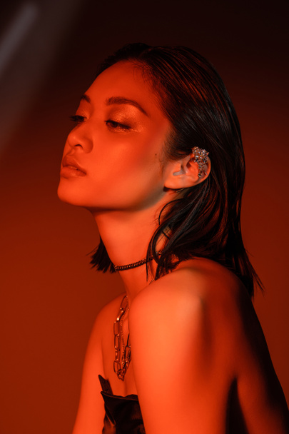 portrait of alluring asian woman with short hair and wet hairstyle posing in strapless dress with trendy cuff earring and necklaces on dark orange background with red lighting, young model  - Photo, Image