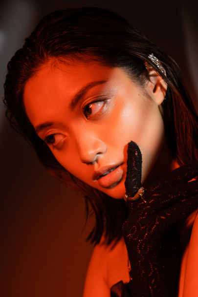 portrait de séduisante femme asiatique aux cheveux courts et coiffure mouillée posant en gant noir avec des anneaux dorés et regardant loin sur fond sombre avec éclairage rouge, jeune modèle, boucle d'oreille manchette  - Photo, image