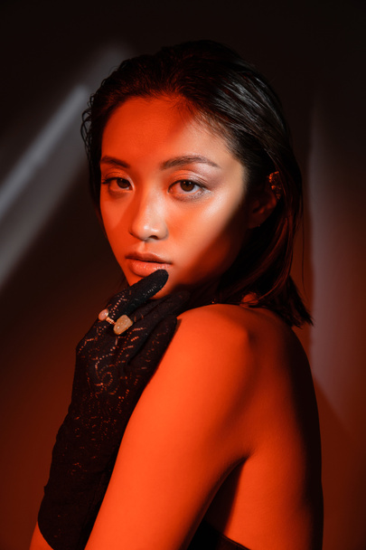 ritratto di giovane donna asiatica con i capelli corti e l'acconciatura bagnata posa in guanto nero con anelli d'oro e guardando la fotocamera su sfondo scuro con illuminazione rossa, modello, bracciale orecchino  - Foto, immagini
