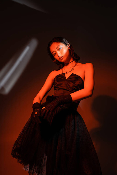 captivating asian woman with wet hairstyle posing in strapless dress with tulle skirt and black gloves with rings while standing on dark orange background with red lighting, looking at camera  - Photo, Image