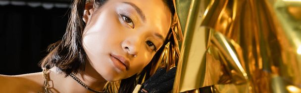 portré csábító ázsiai fiatal nő nedves rövid haj és ékszerek pózol mellett fényes arany háttér, modell, nézi a kamera, ráncos fólia, természetes ázsiai szépség, banner  - Fotó, kép