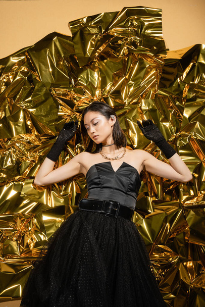 elegant asian young woman with wet hairstyle and short hair posing in black strapless dress with tulle skirt and gloves while standing next to shiny background, model, wrinkled golden foil - Foto, Bild