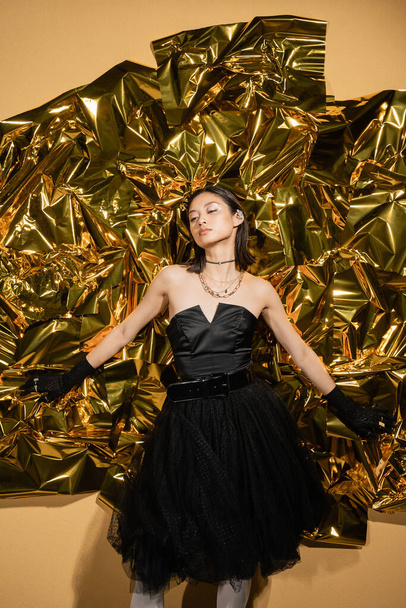 enchanting asian young woman with short hair posing in black strapless dress with tulle skirt and gloves while standing next to shiny yellow background, model, wrinkled golden foil, looking away - Photo, Image