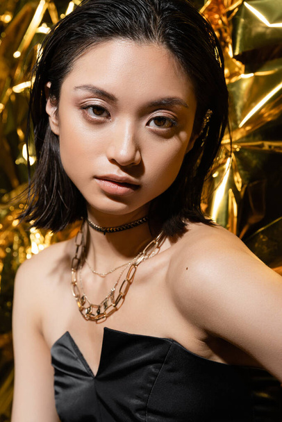 beautiful asian young woman with wet hairstyle and short hair posing in strapless dress with black glove while standing next to shiny golden background, model, looking at camera, wrinkled golden foil - Foto, immagini