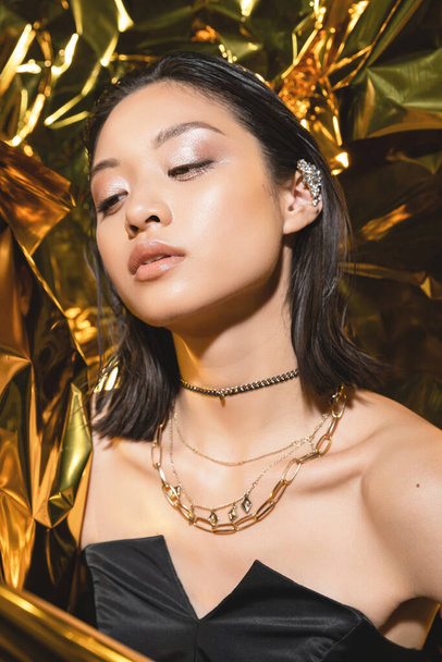 asian young woman with short hair posing in strapless dress with black glove while standing next to shiny yellow background, model, looking away, wrinkled golden foil, wet hairstyle  - Foto, Imagem