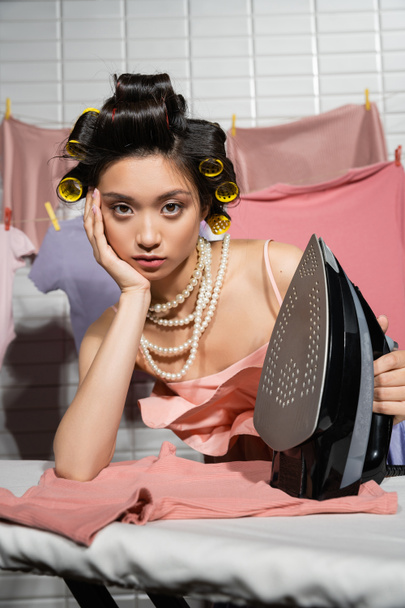 znudzona azjatycka gospodyni domowa z lokówkami do włosów trzymającymi żelazo podczas pozowania w różowym naszyjniku i perłowym naszyjniku w pobliżu czystych i mokrych ubrań wiszących na rozmytym tle, prace domowe, młoda kobieta, pranie  - Zdjęcie, obraz