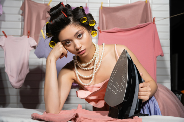 tired asian housewife with hair curlers in pink ruffled top and pearl necklace looking at camera while holding iron near clean clothes hanging on blurred background, housework, young woman, laundry  - Foto, afbeelding