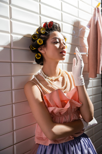 asian young woman with hair curlers standing in pink ruffled top, pearl necklace and gloves while holding cigarette near wet laundry hanging near white tiles, housewife, looking away, smoking  - 写真・画像