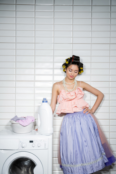 azjatycka młoda kobieta z lokami do włosów stojąca z ręką na biodrze w falbankach, perłowy naszyjnik i spódnica tiulu w pobliżu miski myjącej brudne ubrania na nowoczesnej pralce z detergentem w pralni  - Zdjęcie, obraz