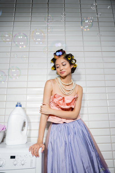 jolie asiatique jeune femme avec cheveux bigoudis debout dans volants top, collier de perles et jupe en tulle près de moderne machine à laver avec détergent dans la buanderie, femme au foyer, regarder ailleurs, bulles de savon - Photo, image