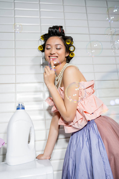 šťastný a mladý asijský žena s vlasy natáčky stojící v načechrané horní, perlový náhrdelník a tyl sukně v blízkosti moderní pračky s čisticí láhve v prádelně, hospodyňky, mýdlové bubliny - Fotografie, Obrázek