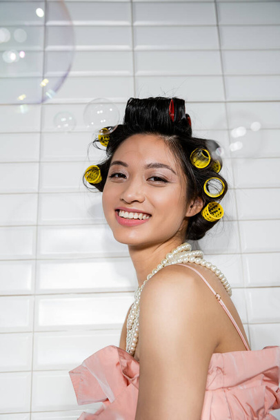 portrait de joyeuse et jeune femme asiatique avec des boucles de cheveux debout dans collier de perles près de bulles de savon floues dans la buanderie avec des carreaux blancs, femme au foyer, beauté naturelle  - Photo, image