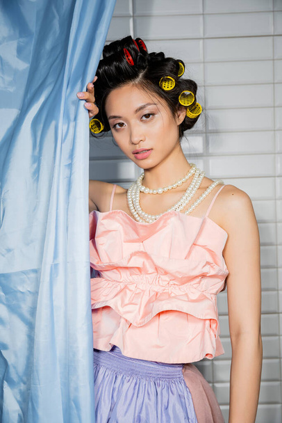 morena e asiática jovem mulher com encrespadores de cabelo de pé em rosa babados topo com colar de pérolas perto de cortina de banheiro azul e olhando para a câmera perto de azulejos brancos em casa  - Foto, Imagem