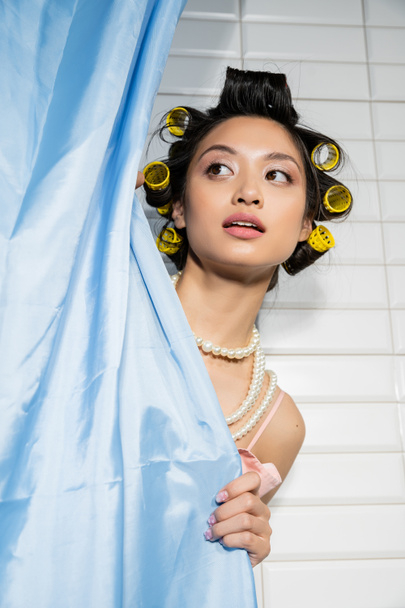 curious and asian young woman with hair curlers standing in pearl necklace behind blue bathroom curtain and looking away near white tiles at home, housewife, domestic scene  - Foto, Imagem