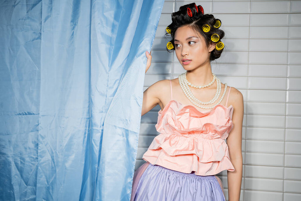 alluring and asian young woman with hair curlers standing in pink ruffled top with pearl necklace near blue bathroom curtain and looking away near white tiles at home  - Photo, Image