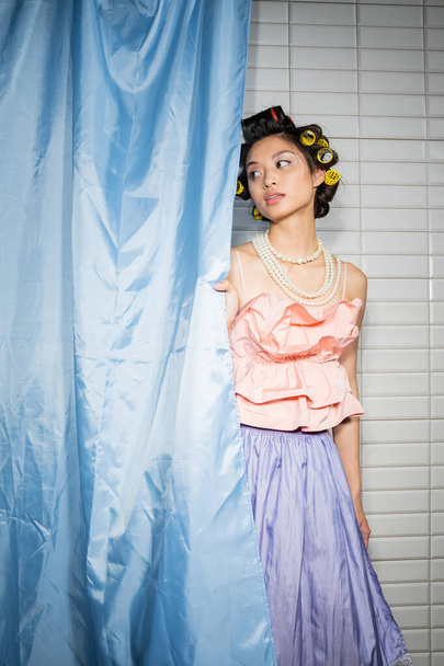 brune et asiatique jeune femme avec des bigoudis de cheveux debout en haut à volants rose avec collier de perles et jupe près rideau de salle de bains bleu et regarder loin près de tuiles blanches à la maison  - Photo, image