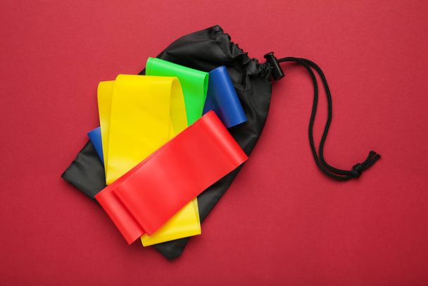 A set of colorful latex rubber bands for fitness and a black cover for them on red background. Sports concept. Top view - Φωτογραφία, εικόνα