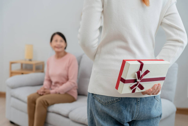 Asyalı genç kızlar anneleri evde kanepede otururken anneleri için hediye kutuları saklıyorlar. Evde özel bir gün kutluyoruz. Mutlu yıllar sana. - Fotoğraf, Görsel