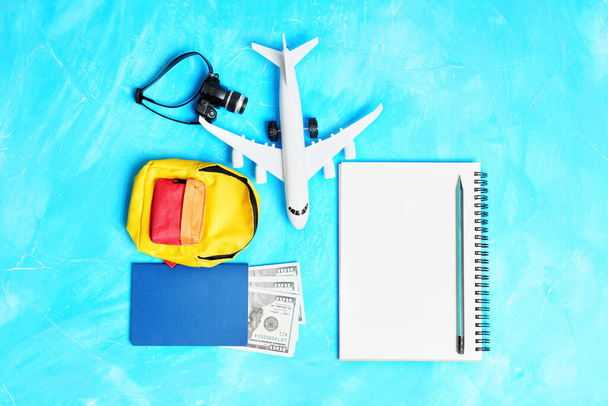 The concept of getting ready for a plane trip: Toy plane model, notebook with a pencil, vibrant miniature backpack, passport with cash and tiny camera arranged on a blue background. - Photo, Image