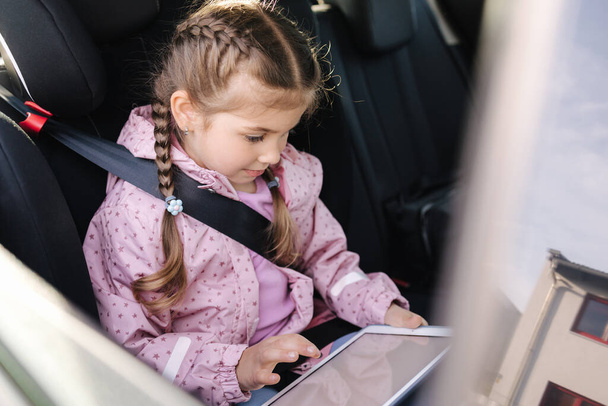 Belle petite fille dans le siège auto en utilisant une tablette numérique pendant le voyage. Joyeux enfant femelle sur le siège arrière. Photo de haute qualité - Photo, image