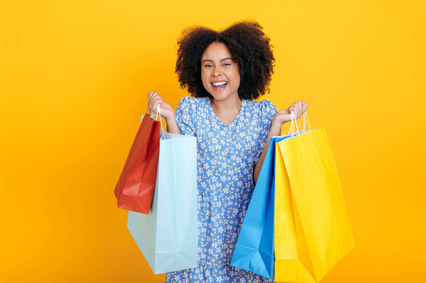 Joyful beautiful trendy curly haired hispanic or brazilian woman wearing blue sundress, holding paper shopping bags, looks at camera and smiling happily, stand on isolated yellow background - Фото, изображение