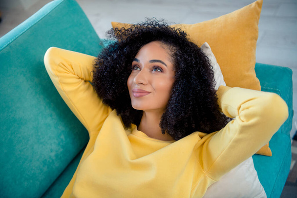 Портрет мрійливої розслабленої мирної дівчини з хвилястою зачіскою, одягненою в жовтий стильний светр, лежав на подушках за головою в приміщенні
. - Фото, зображення