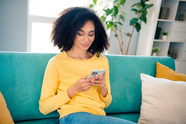 Portrait of gorgeous clever lovely girl with wavy hairstyle dressed yellow sweatshirt texting email on smartphone sit on sofa indoors room. - Foto, Imagen
