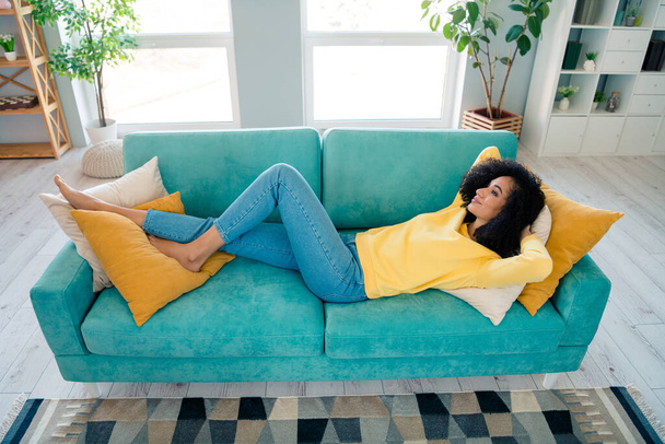 Portrait of positive relaxed lovely girl with wavy hairstyle dressed yellow sweatshirt lying on couch arms behind head indoors room. - Foto, Imagen