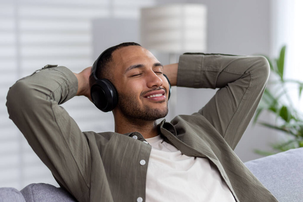 Young african american man relaxing at home wearing headphones. Listens to music, podcast, audiobook. Relaxed, he sits on the sofa, threw his arm behind his head, closed his eyes. - Valokuva, kuva