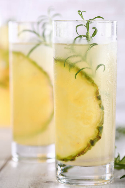 A glass of lemonade with pineapple pieces, ice cubes and rosemary. Cold refreshment organic non-alcohol cocktail. - Foto, imagen