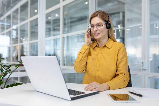 Portrait of a young woman in a headset working in an office center online from a laptop. - Фото, изображение