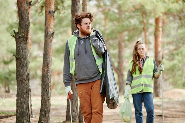 Voluntario frunciendo el ceño llevando bolsa de basura con basura recolectada en el bosque - Foto, imagen