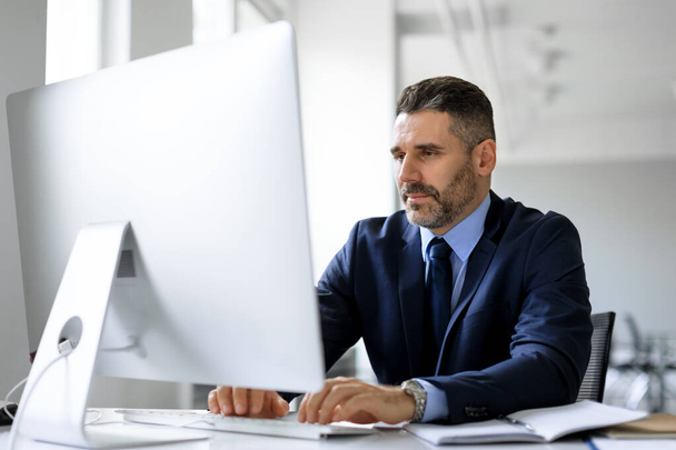 Homme d'affaires d'âge moyen en costume assis à la table de travail au bureau devant l'ordinateur, tapant sur le clavier, envoyant des e-mails à ses partenaires d'affaires, travaillant sur la recherche marketing, l'espace de copie - Photo, image