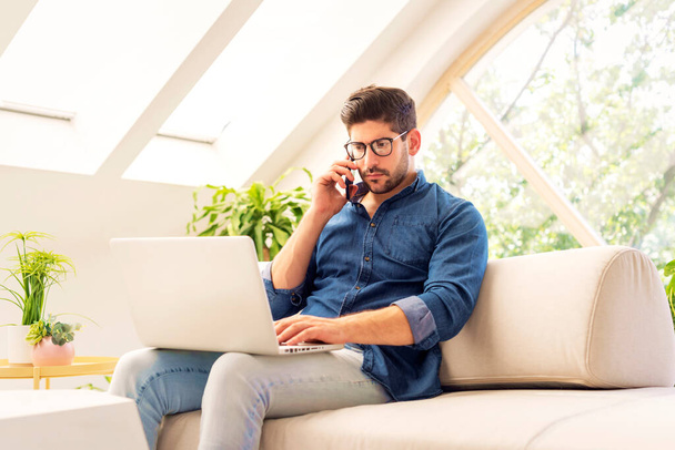 Thinking man using smartphone and laptop while working from home. Confident male sitting on the couch and having a phone call. Home office. - Foto, Imagen
