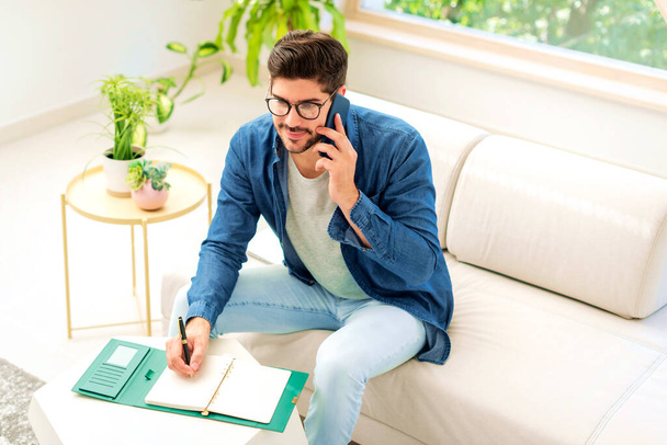 Portrait shot of happy businessman using phone and talking with somebody while sitting on sofa and working from home. Confident man taking notes on notepad. - Zdjęcie, obraz