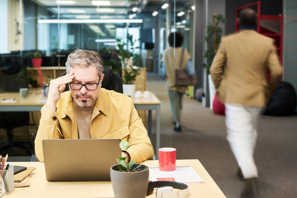 Seriöser Analyst oder Manager analysiert Online-Daten, während er am Arbeitsplatz vor dem Laptop sitzt und seine Kollegen an ihm vorbeigehen - Foto, Bild