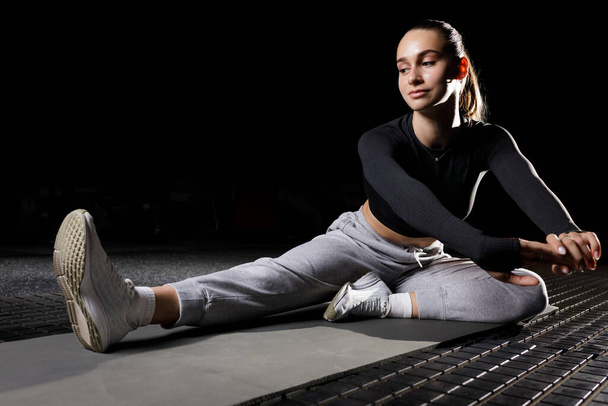 Mladá fitness dívka protahuje nohy na podložce v tělocvičně izolované na černém pozadí. Sportovní žena se zahřívá na podlaze - Fotografie, Obrázek