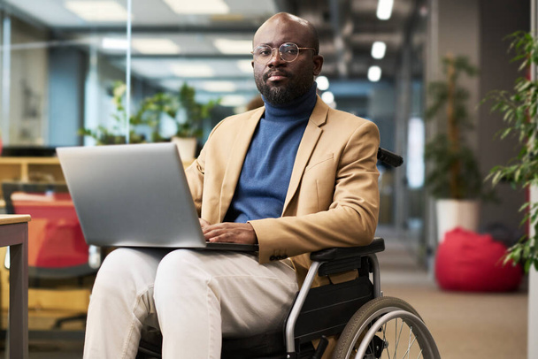 Dizlerinin üzerinde diz çöküp, uzay ofisindeki tekerlekli sandalyede oturan ve kameraya bakan genç ve ciddi bir Afrikalı Amerikalı iş adamı. - Fotoğraf, Görsel