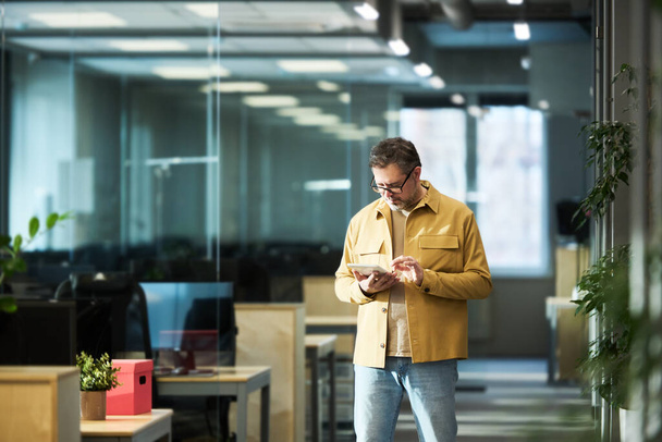 Corredor masculino maduro en atuendo casual usando tableta mientras está de pie en el pasillo en la oficina de espacio abierto y mirando a través de datos financieros en línea - Foto, imagen