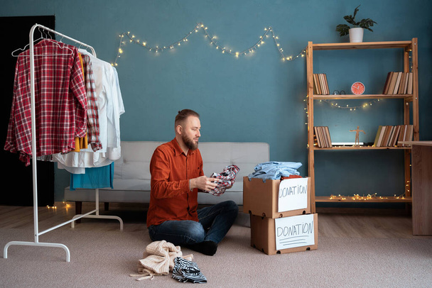 Ένας άντρας βάζει ρούχα σε κουτί δωρεών στο σπίτι. Η ιδέα της δωρεάς. Αντιγραφή χώρου - Φωτογραφία, εικόνα