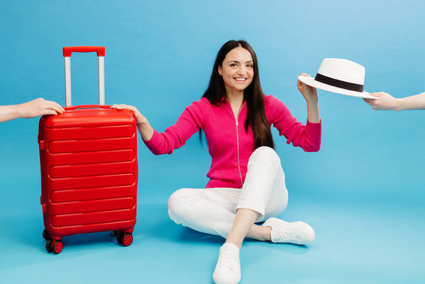 Happy young tourist woman sitting at the suitcase and going to travel on holidays isolated on blue background - Photo, Image